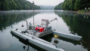 A grey twin-hull survey USV on a misty reservoir lowers a yellow open-frame ROV from a davit while a water sampling rack sits on its deck